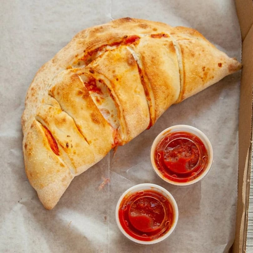A golden-brown calzone with visible cheese and sauce, served with two cups of marinara.