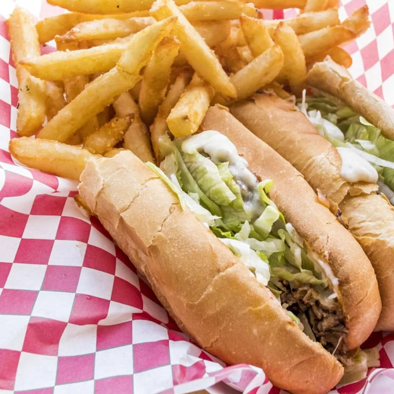 Two Basil's cheesesteak subs with lettuce and sauce, alongside a pile of golden french fries.