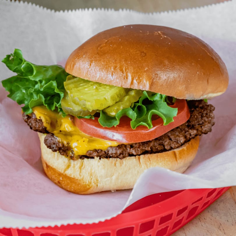 A Basil's cheeseburger with lettuce, tomato, and pickles in a red basket.